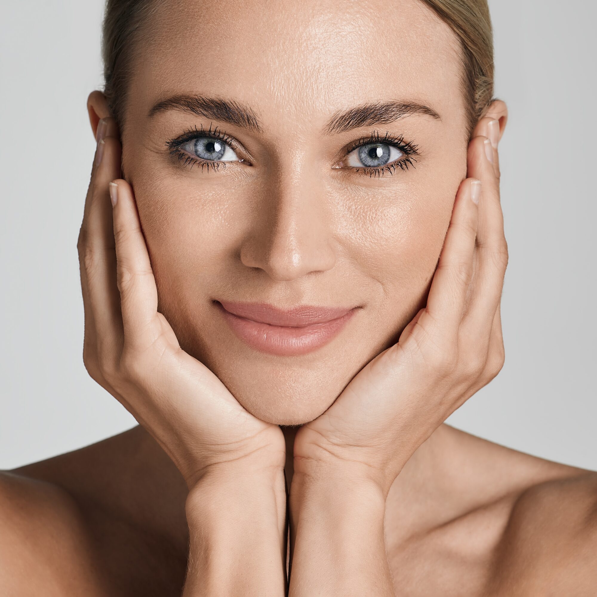 Beautiful woman with healthy facial skin touching hands moisturized face skin, on light grey background.